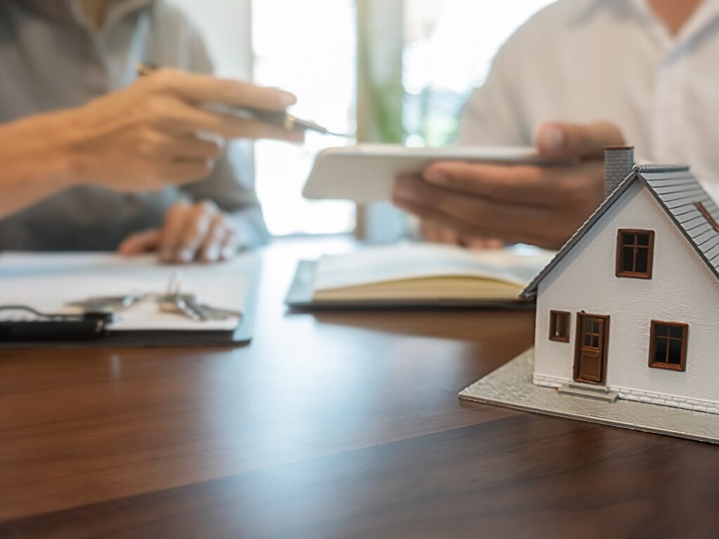 A close-up of a miniature house on a desk while two people review paperwork and a tablet in the background—perfect for anyone looking to buy a home in San Diego.